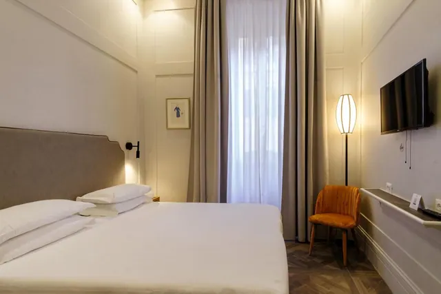 A bedroom with a double bed, a wall-mounted flat-screen television, a floor lamp and an orange upholstered chair beside a window with sheer and taupe curtains.