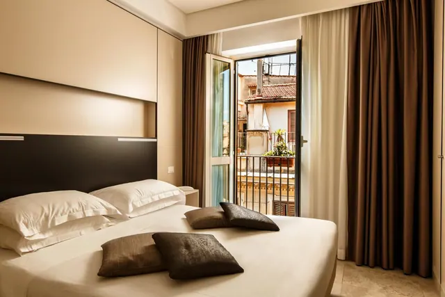 A double bed with white pillows and dark cushions in a bedroom, with open French doors onto a balcony overlooking older buildings with terracotta roofs.
