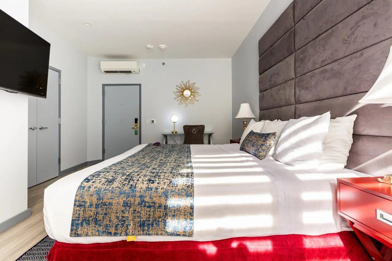 A hotel room with a king bed under a patterned blue and gold runner, a tufted grey headboard, a red nightstand, a wall-mounted television and a desk with a chair.