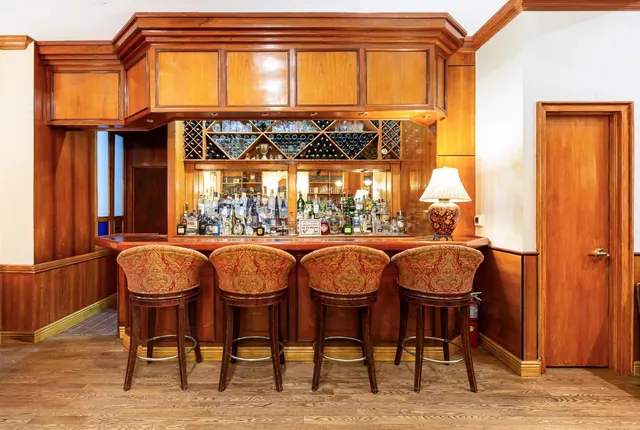 A wood-panelled bar with four upholstered swivel stools in front of shelves of liquor bottles and a wine rack.