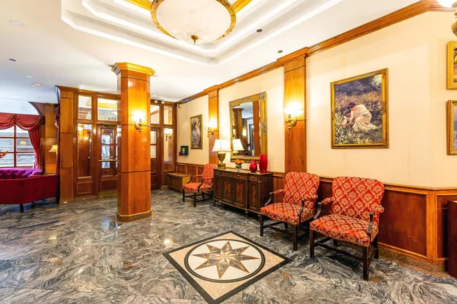 The lobby has a patterned marble floor with a star medallion, red upholstered armchairs, a wooden sideboard, a mirror and framed paintings on the walls.