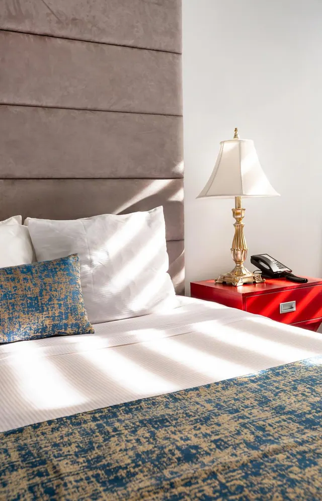 A made bed with white linen and a blue and gold throw sits beside a red nightstand holding a gold table lamp and a telephone, with sunlight falling across the bedding.