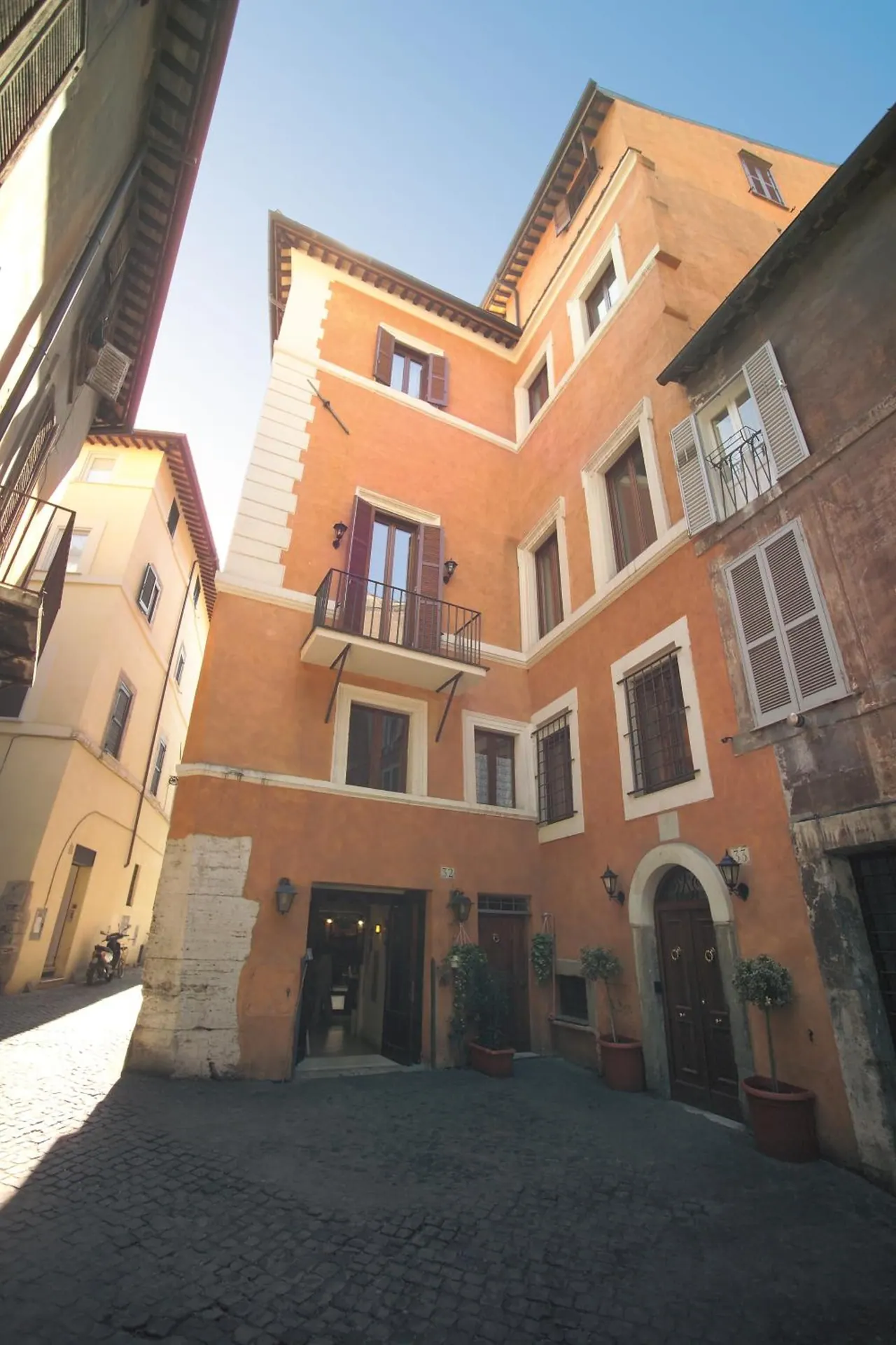 Wide-angle view of an orange multi-storey building with shuttered windows, a small balcony and a doorway onto a cobbled street under a clear blue sky.