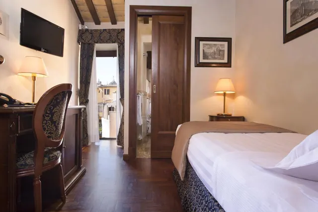 A hotel bedroom with a single bed, a wall-mounted television, a wooden desk and chair, and a view through an open doorway to rooftops and buildings outside.