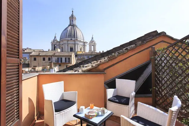 A small terrace with white wicker chairs and a glass table, with a view over rooftops to a domed church.