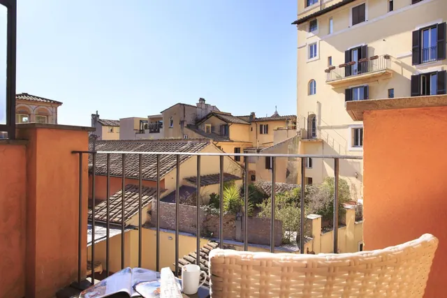 A wicker chair and a table holding a mug and a magazine sit on a balcony overlooking rooftops and yellow and orange buildings under a clear blue sky.