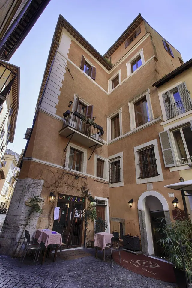 Exterior of a multi-storey ochre building on a narrow cobbled street, with a balcony above, outdoor tables with checked cloths, and an arched entrance with a red carpet.