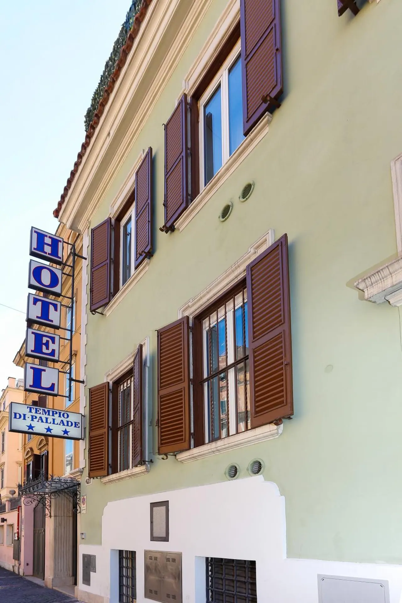 The street-facing exterior of a pale green hotel building with brown shutters, a hanging HOTEL sign and a Tempio di Pallade sign.