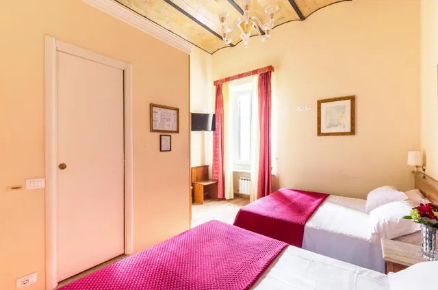 A twin-bedded hotel room with pink bedspreads, a vaulted ceiling, a glass chandelier, and a window with red curtains.