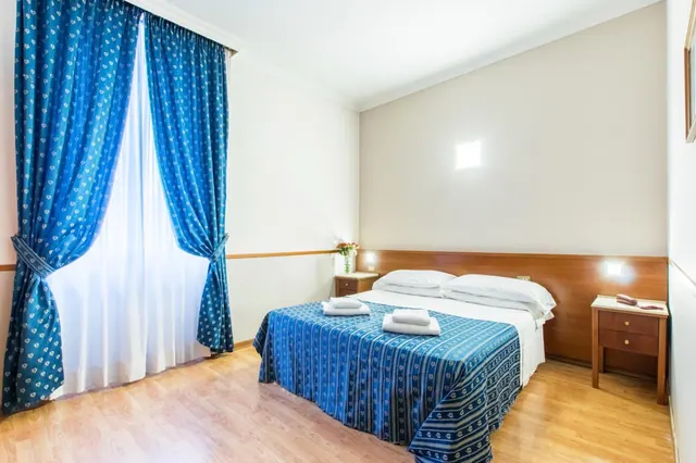 A bedroom with a double bed under a blue striped bedspread, blue patterned curtains at the window, a wooden headboard and two wooden bedside tables.