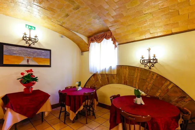 A dining room with a brick vaulted ceiling, several tables with red tablecloths, wall sconces, a framed painting and a small window with a curtain.