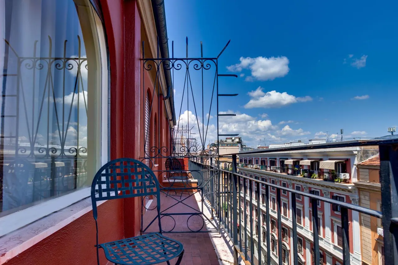 A tiled balcony with two wrought iron chairs beside a red wall and an arched window, with neighbouring apartment buildings and blue sky beyond the railing.
