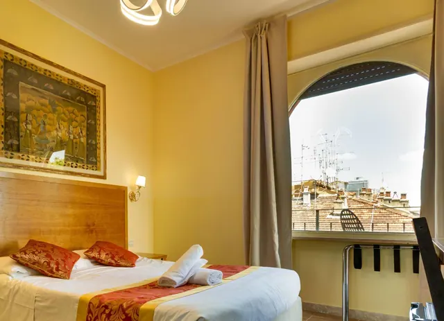 A double bed with red patterned cushions and folded white towels sits beside an arched window looking out over terracotta rooftops and TV antennas.