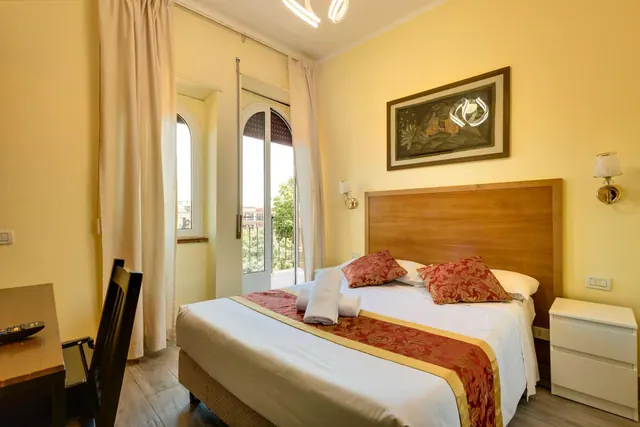 A double bed with a red and gold runner and cushions sits in a yellow bedroom, with a desk and chair on the left and an open balcony door showing trees and buildings outside.