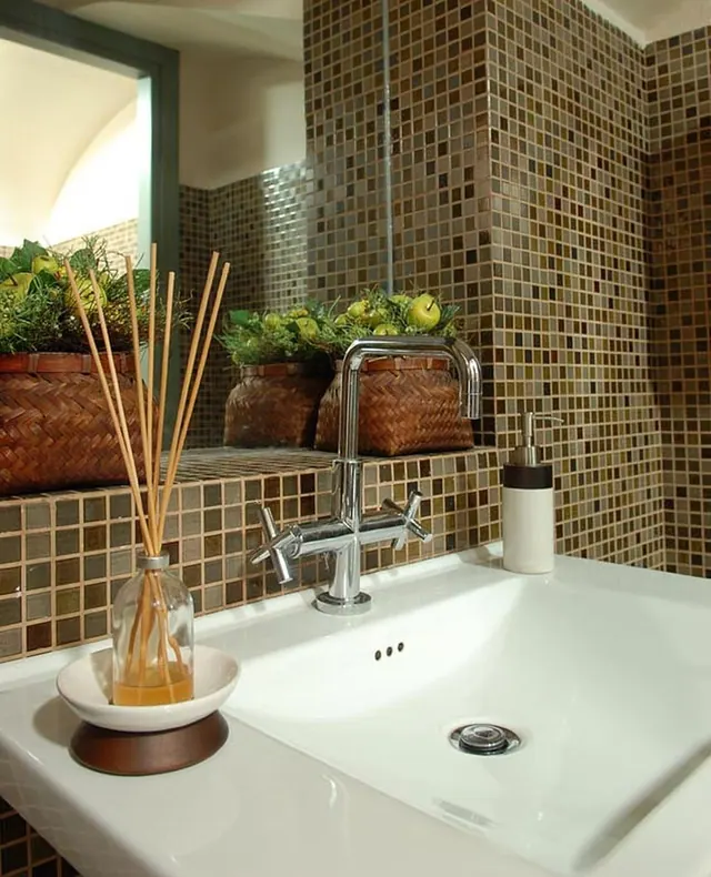 A white basin with a chrome tap, a reed diffuser and a soap dispenser sits in front of mosaic tiled walls and woven baskets of greenery and apples.