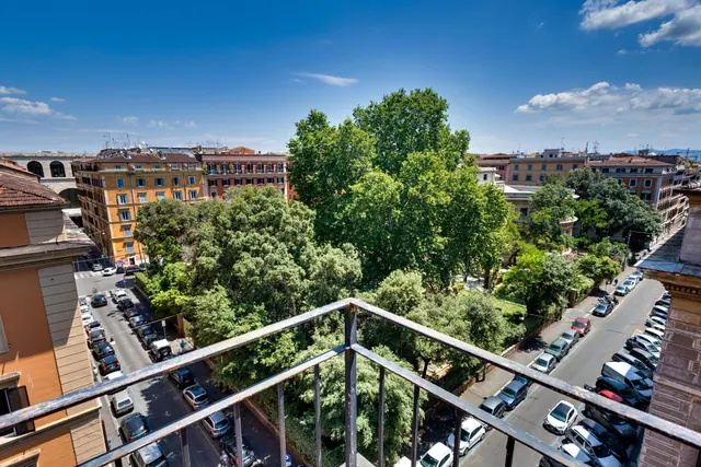 View from a balcony with a metal railing over tree-covered gardens, parked cars on a street, and ochre-facaded apartment buildings under a blue sky.