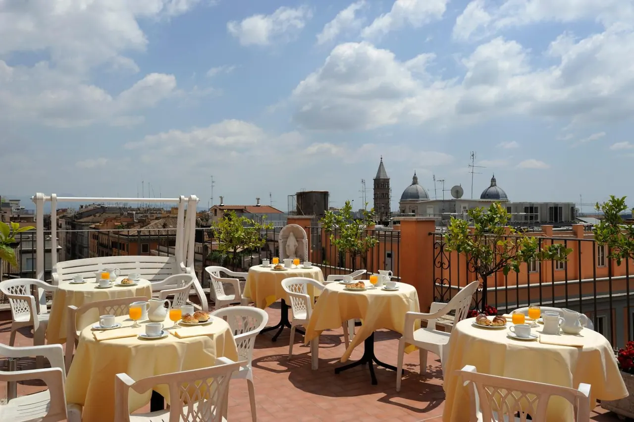 A rooftop terrace with white chairs and tables set with yellow cloths, cups, orange juice and pastries, with domed churches and rooftops in the background.