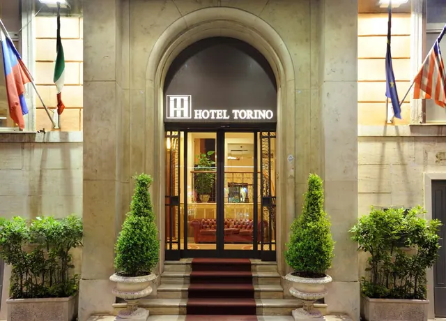 Arched stone entrance of Hotel Torino at night, with an illuminated sign, clipped topiary in urns, a red-carpeted staircase and flags on the facade.
