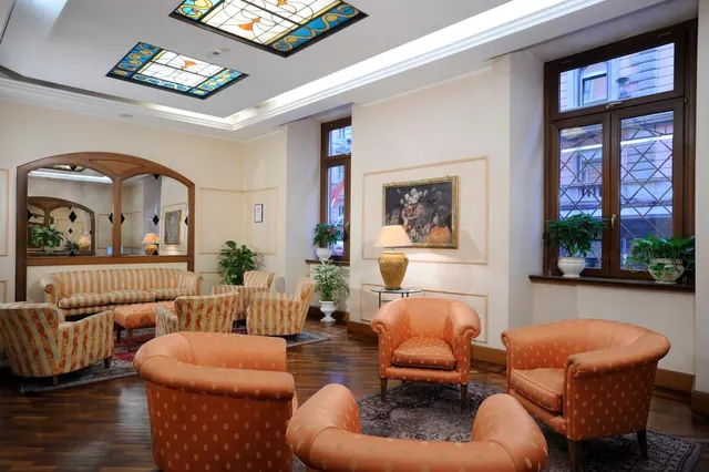 A hotel lobby lounge with orange upholstered armchairs, a striped sofa, stained glass ceiling panels, and windows looking onto a building across the street.