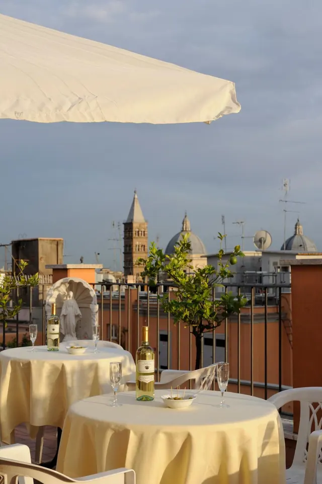 Rooftop terrace with cream-clothed tables, wine bottles and glasses, a statue in a niche, and a view over rooftops and church towers under a cloudy sky.