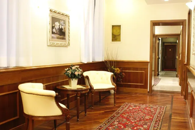 A seating area with two cream armchairs, a round wooden side table with a flower vase, wood wall panelling and a red patterned rug on a wood floor, with a doorway leading to a corridor.