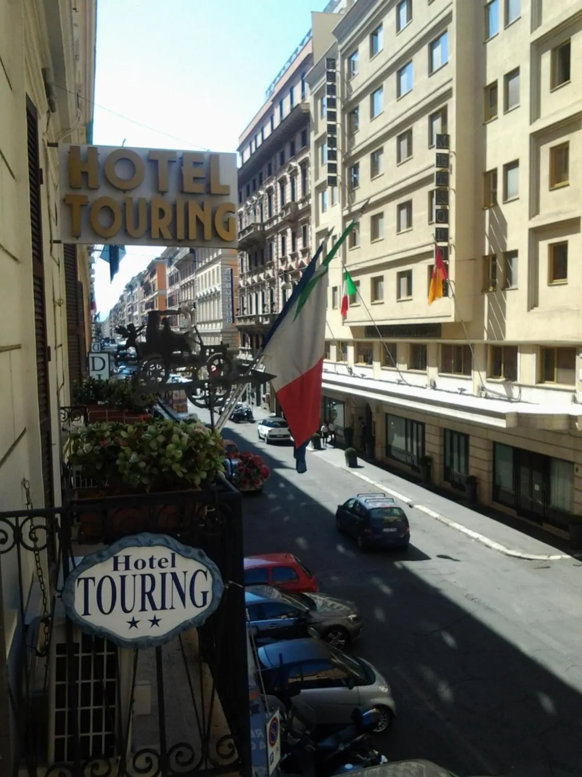 A view from a wrought iron balcony with plants down a sunlit Rome street lined with parked cars, cream-coloured hotel buildings and Hotel Touring signs, with a flag hanging from a building.
