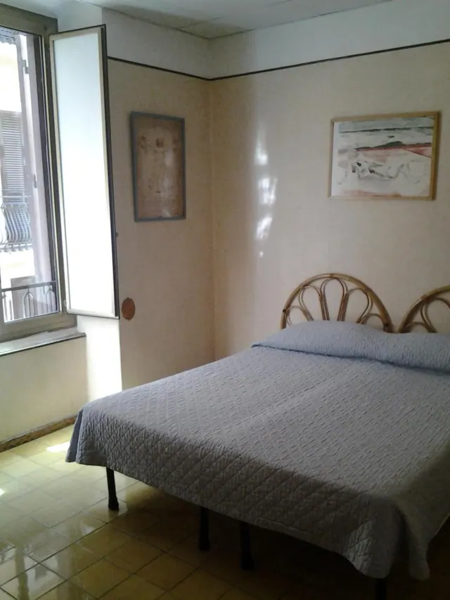 A double bed with a grey quilted cover and rattan headboard stands in a bedroom beside an open window, with two framed pictures on the walls.