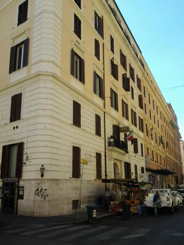 Exterior of a cream-coloured multi-storey hotel building with brown shutters on a Rome street, with a fruit stall and a parked white pickup truck at street level.