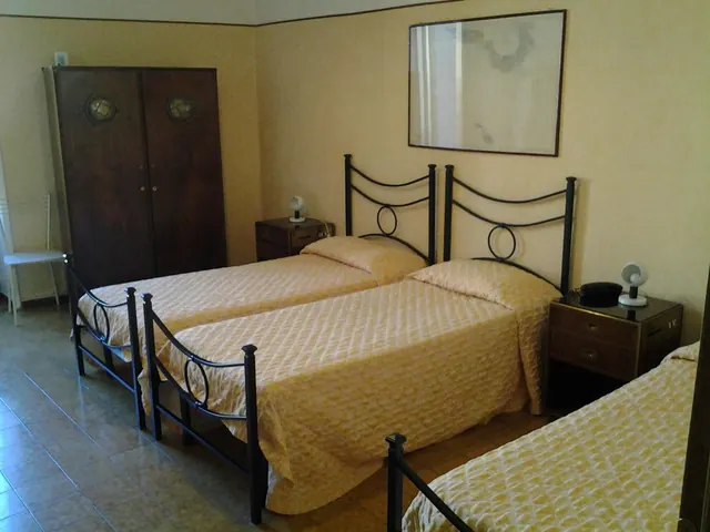 A bedroom with three single beds with metal headboards, a wooden wardrobe, two bedside tables with lamps, and a framed picture on the wall.