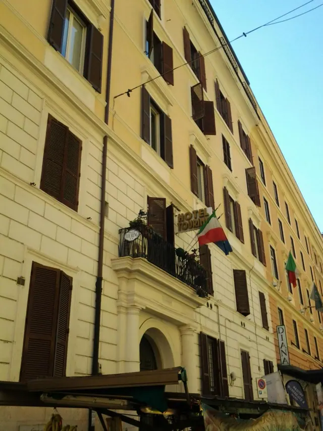 Low-angle street view of a cream-coloured multi-storey building with brown shutters, a small railed balcony with plants, a hotel sign, and Italian flags hanging on the facade.