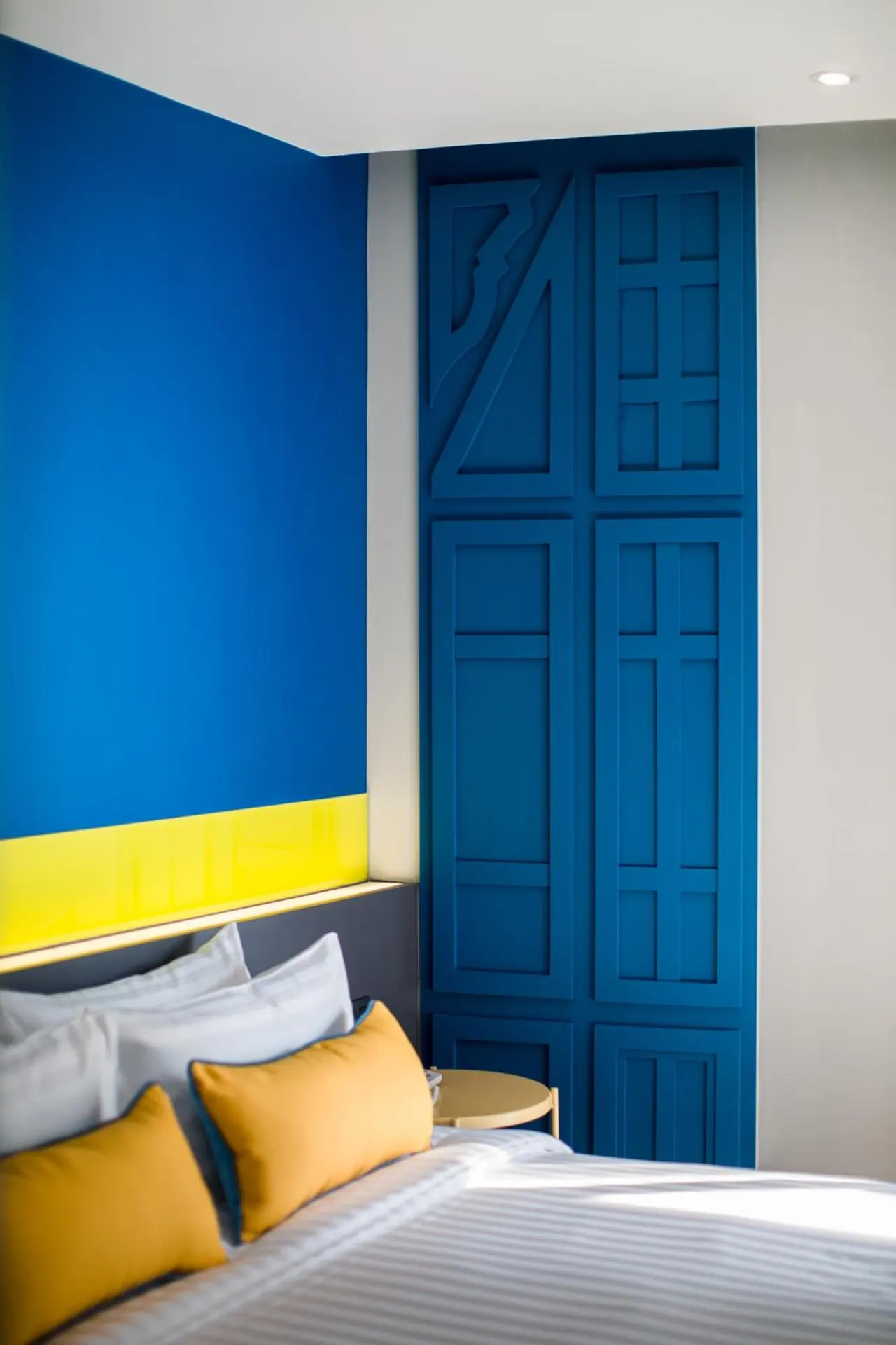A bedroom with bright blue walls, a yellow wall band, a blue paneled wall, and a bed with white and yellow cushions.