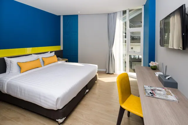 A bedroom with a king bed with yellow cushions, a yellow chair at a wooden desk, a wall-mounted television and floor-to-ceiling windows looking onto a neighbouring building.