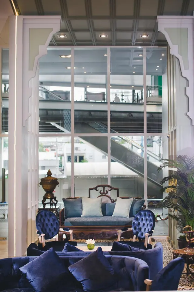 A lounge area with blue velvet sofas and tufted armchairs, a carved wooden settee, and large windows looking onto an escalator and walkway outside.