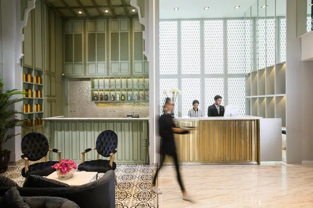 A hotel lobby with a reception desk where two staff stand, a blurred figure walking past, dark velvet armchairs and a patterned tile floor.