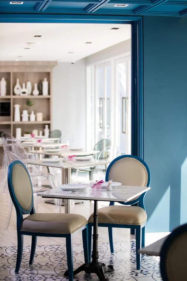 A restaurant dining room with blue-framed chairs and white marble-top tables on a patterned tile floor, with shelves of white vases and tall windows in the background.