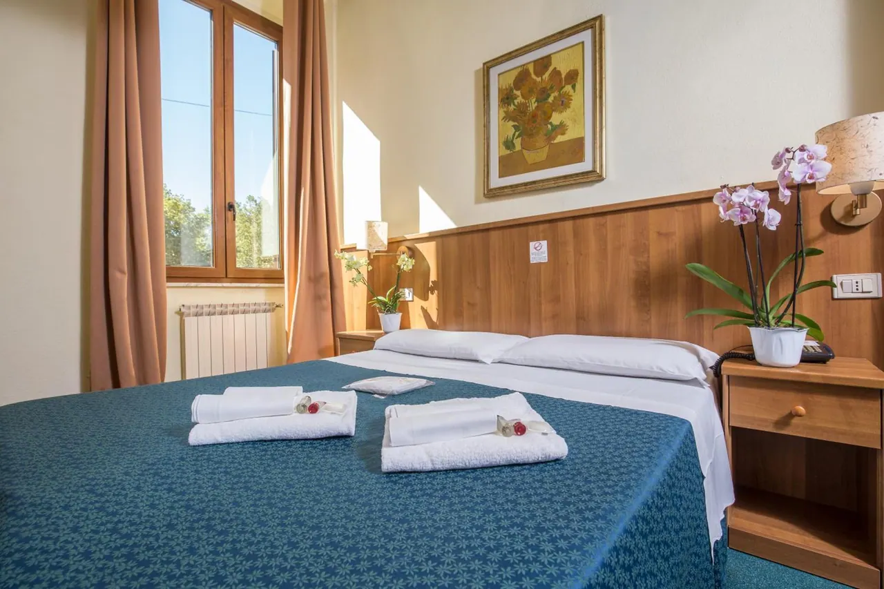 A double bed with a blue bedspread and folded white towels in a hotel bedroom, with a window showing trees outside, a framed painting and a potted orchid on the nightstand.
