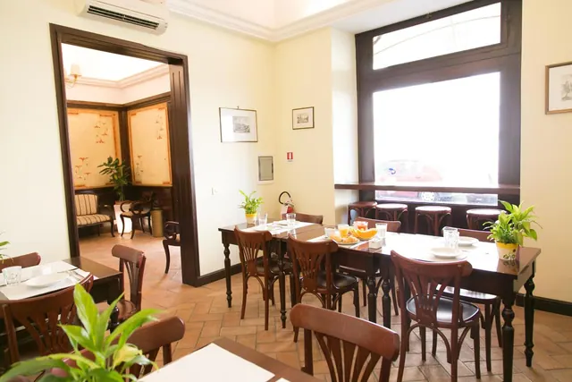 A breakfast room with set wooden tables and chairs, a large window, a wall-mounted air conditioner and an adjoining seating area with a striped sofa.