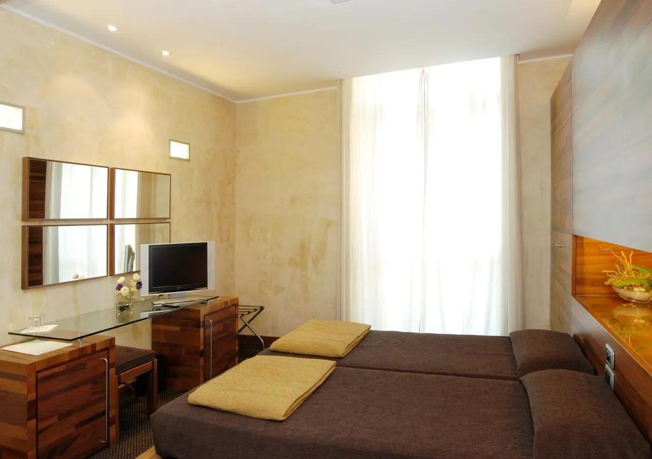 A bedroom with two beds covered in brown throws, a glass-topped desk with a flat-screen television, mirrors on the wall, and tall white curtains over a window.