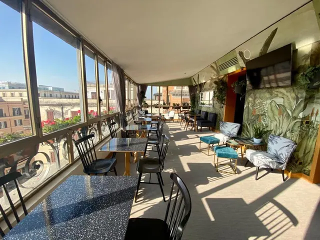 A glass-walled dining area with black chairs and speckled tables looks out over Rome rooftops and buildings, with armchairs and a tropical plant mural on the right.