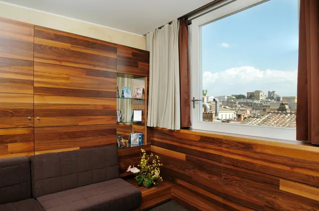 A lounge area with wood-paneled walls, a dark upholstered sofa, a small bookshelf and a window looking out over Rome rooftops and buildings.