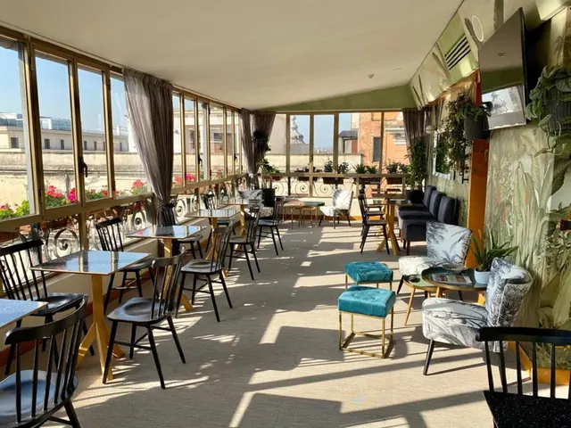 A glass-enclosed terrace dining area with tables, chairs and lounge seating, with windows looking out over Rome rooftops.