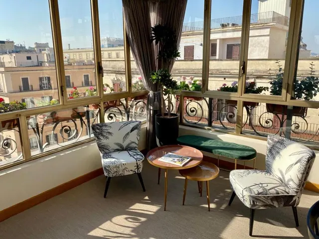 A sitting area with two patterned armchairs, a green bench, round side tables and tall windows looking out over rooftops and buildings.