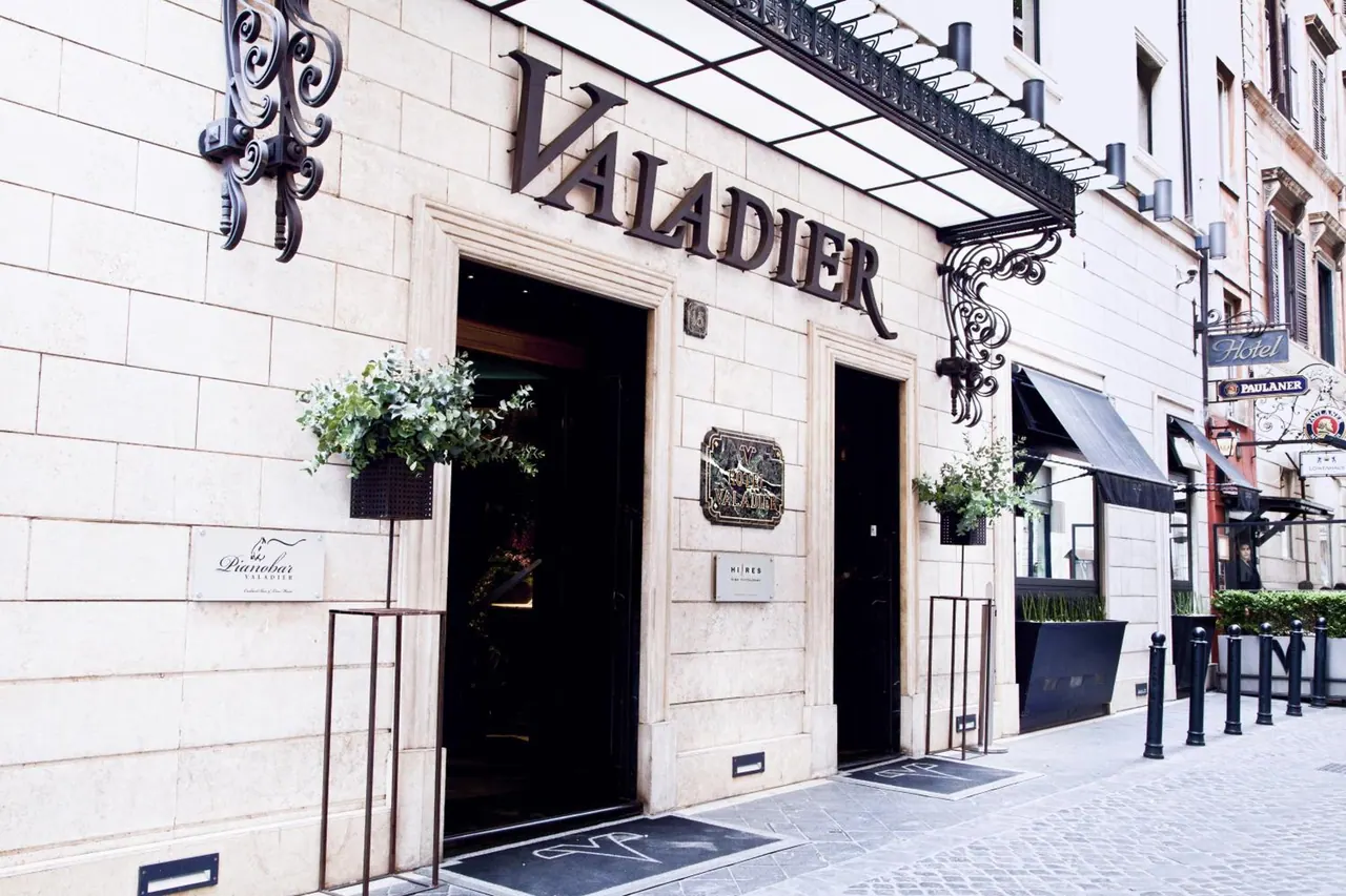 The street facade of the Valadier hotel, with its name in large letters above a glass canopy, a dark doorway, potted plants and a cobbled pavement with bollards in front.