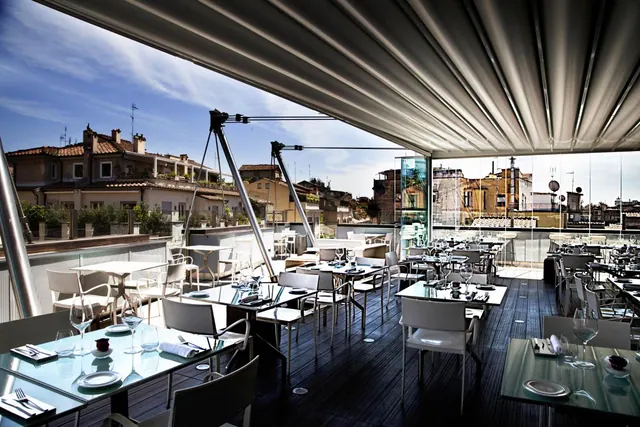 An open rooftop dining terrace under a retractable awning has set tables and white chairs, with terracotta rooftops of the city visible beyond.