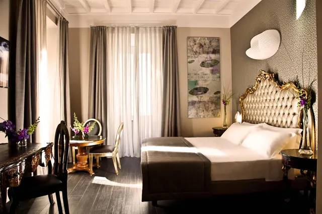 A bedroom with a double bed under an ornate gilded and tufted headboard, a round table with chairs and a dark wooden desk, with sheer curtains over a window.