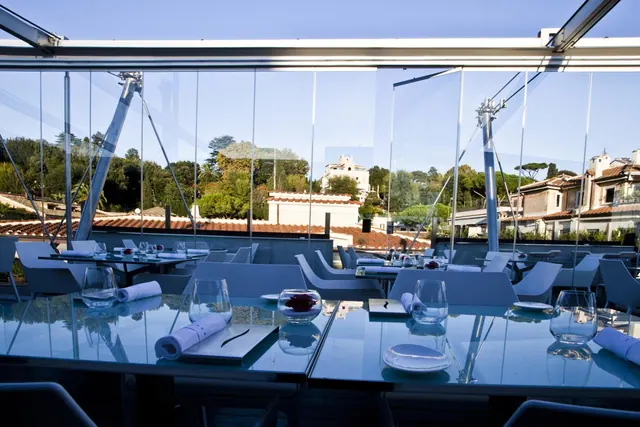 A glass-walled restaurant terrace with set tables and chairs, looking out over rooftops and trees.