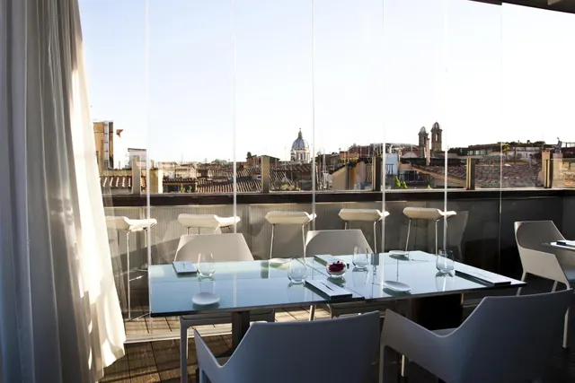 A glass-walled rooftop terrace with a set dining table and white chairs overlooks Rome's rooftops and a domed church.