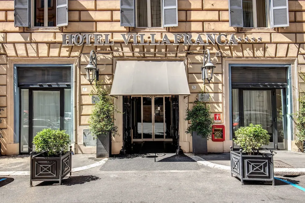 The front facade of Hotel Villa Franca, with a cream awning over the entrance, potted plants beside the doors, and wall lamps on the stone building.