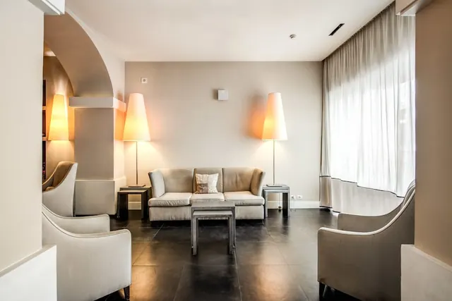 A lounge area with a sofa, armchairs, two floor lamps and a sheer-curtained window, on a dark tiled floor.