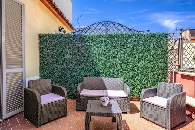 A furnished terrace with two wicker armchairs, a wicker two-seat sofa and a coffee table stands against a green hedge wall under a blue sky.